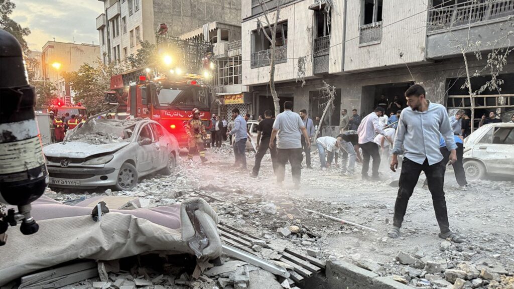 Aerial view of buildings collapsing in a Middle Eastern city following missile strikes, with smoke rising and debris scattered, illustrating the aftermath of a reported U.S. strike on Iran. Image credit: