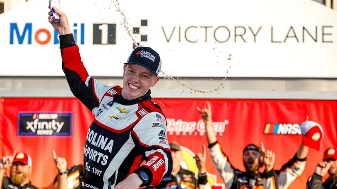 Connor Zilisch celebrating his victory at Watkins Glen, raising his fist in triumph beside his No. 88 JR Motorsports Chevrolet race car.