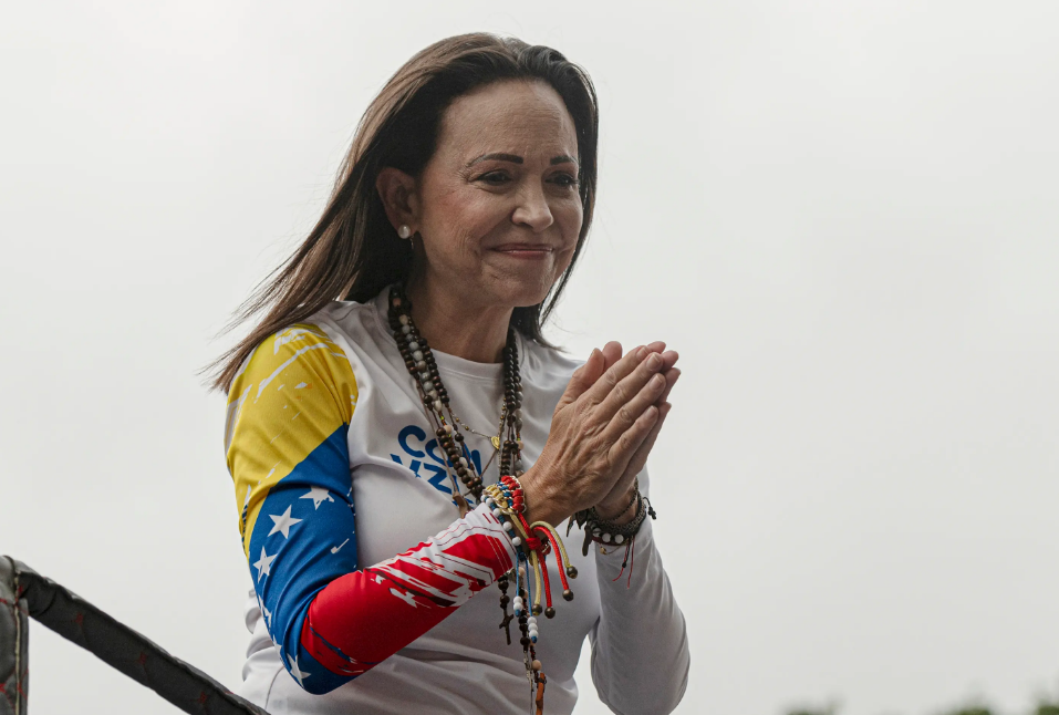 A woman wearing a Venezuela-themed shirt stands with her hands together in a gesture of gratitude during a public appearance.