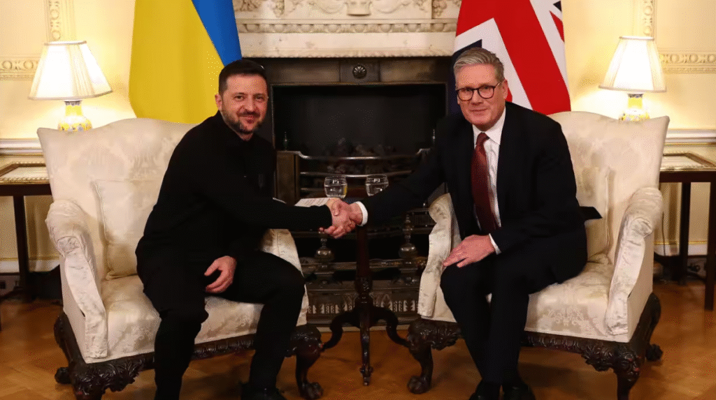Ukrainian President Volodymyr Zelenskyy (left, in black) shakes hands with UK Labour Leader Sir Keir Starmer (right, in a suit) while seated in ornate chairs, with the Ukrainian and Union Jack flags visible behind them.