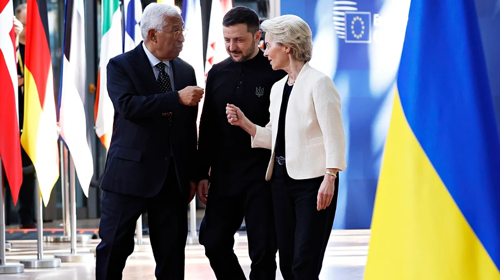 Ukrainian President Volodymyr Zelenskyy (center, in black) walks and gestures alongside António Costa (left, President of the European Council) and Ursula von der Leyen (right, President of the European Commission). The officials are surrounded by EU member state flags and the Ukrainian flag in a European Union setting.